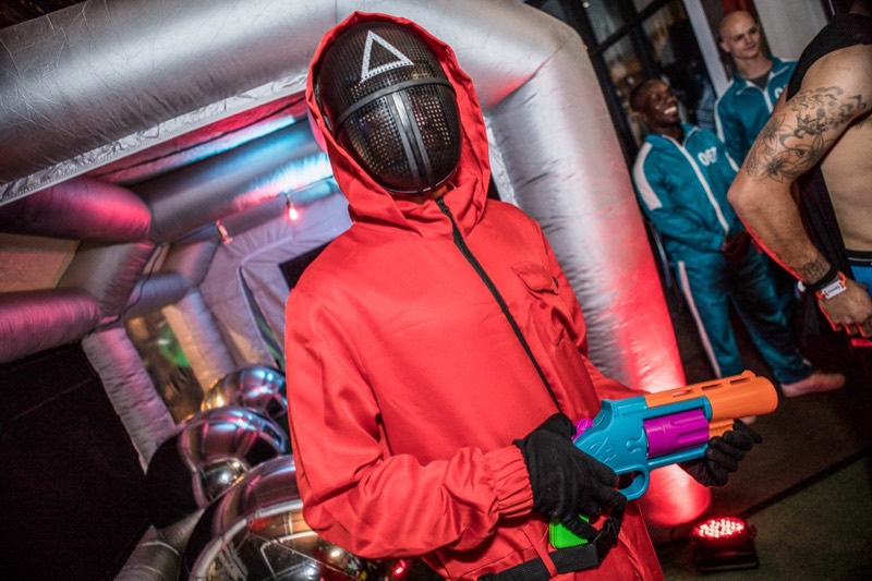 Red jumpsuit guard with a toy blaster, teal tracksuit players behind at Coloween 2021 in Colorado
