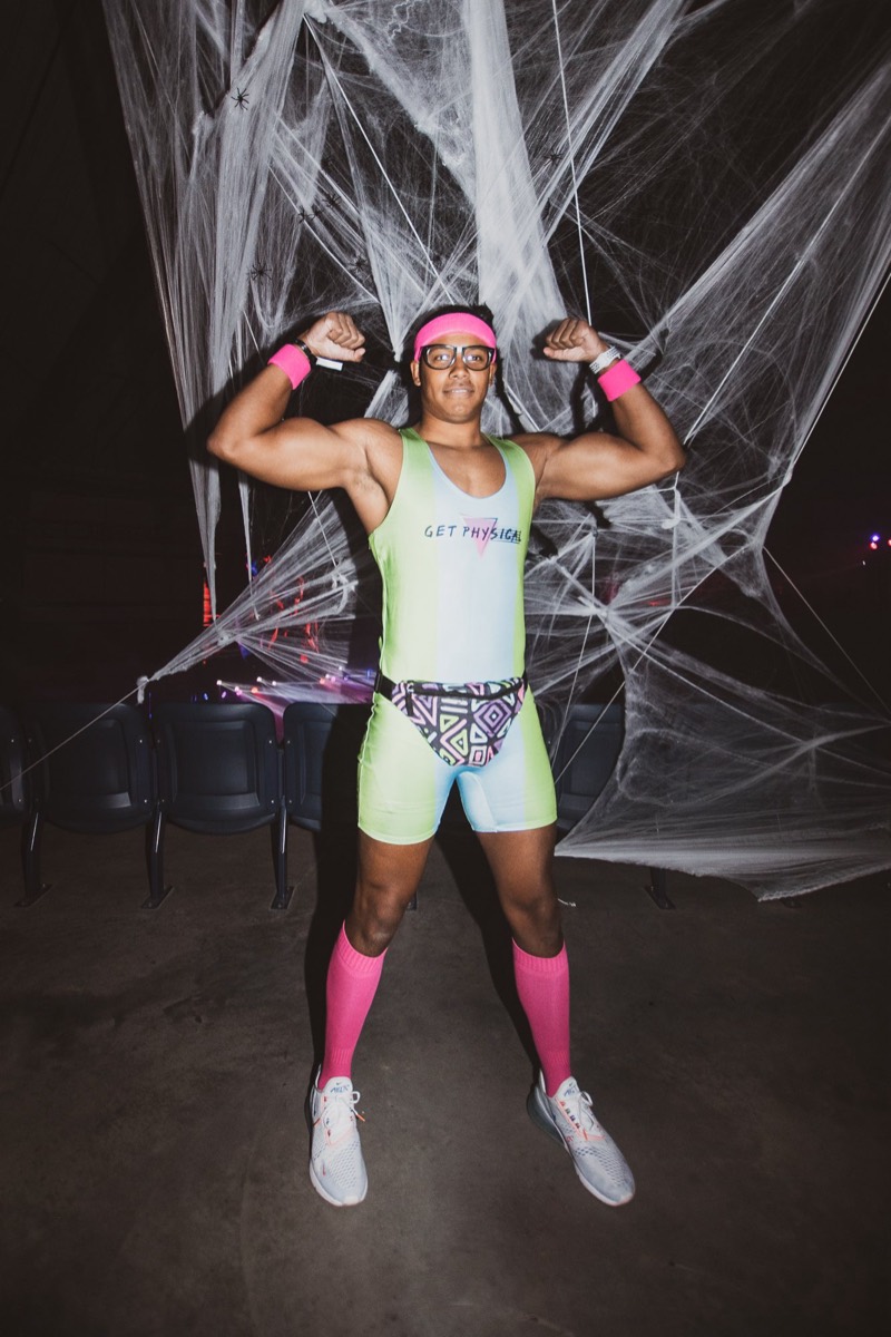 Neon 80s workout guy flexing in front of giant cobwebs at Coloween, Denver's 21+ Halloween party