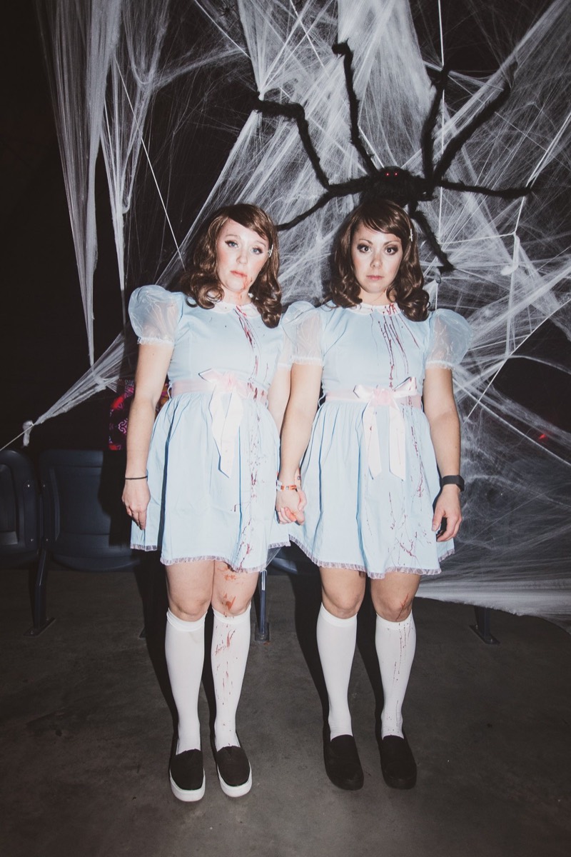 Twin blue dresses holding hands beneath a giant spider web at the adults-only Coloween Halloween event