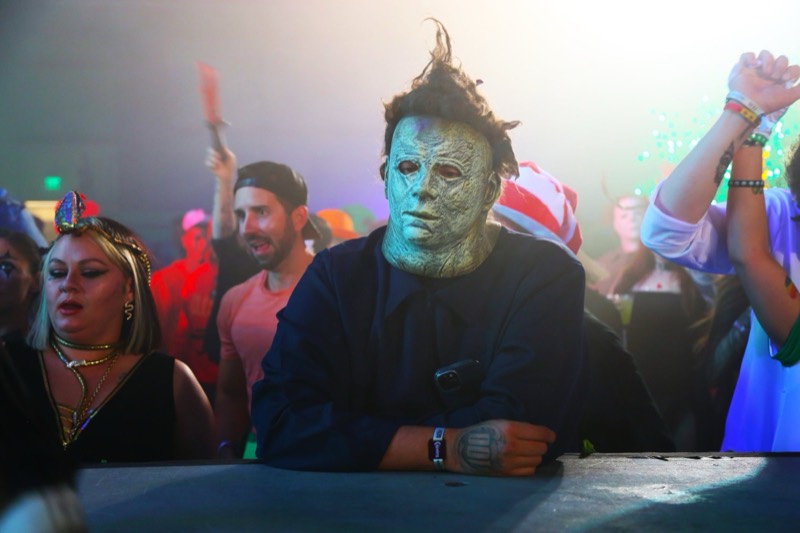 Masked slasher leans on the rail while the crowd dances behind him at the adults-only Coloween Halloween event