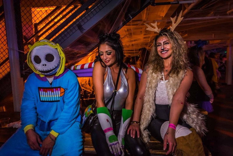 Skeleton onesie and deer-makeup friends laughing together on a bench at Coloween, Denver's 21+ Halloween party