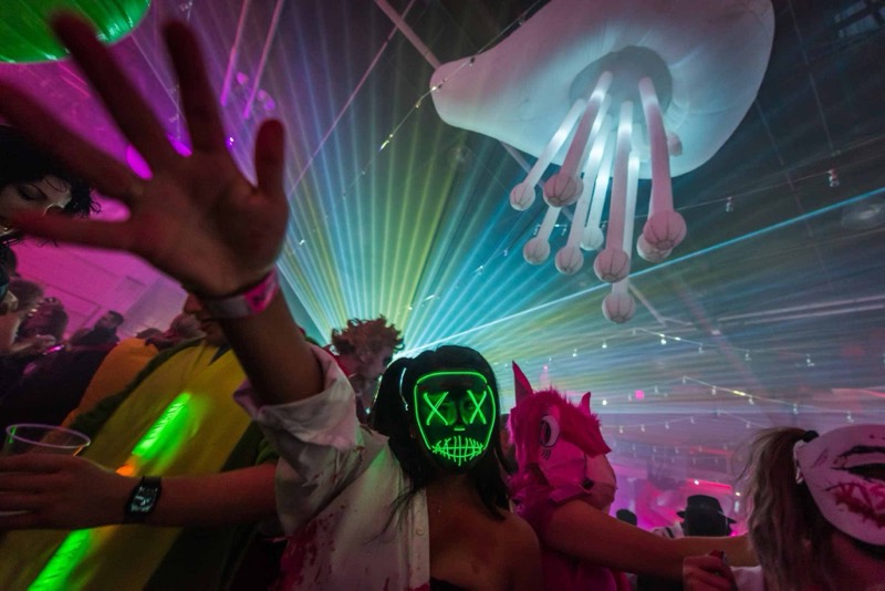 Neon green mask glowing beneath the jellyfish chandelier and lasers at Denver's Coloween Halloween party