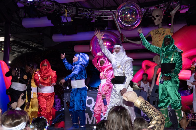 The dance crew hits arms-up in rainbow metallic suits on the dance floor at Coloween 2025